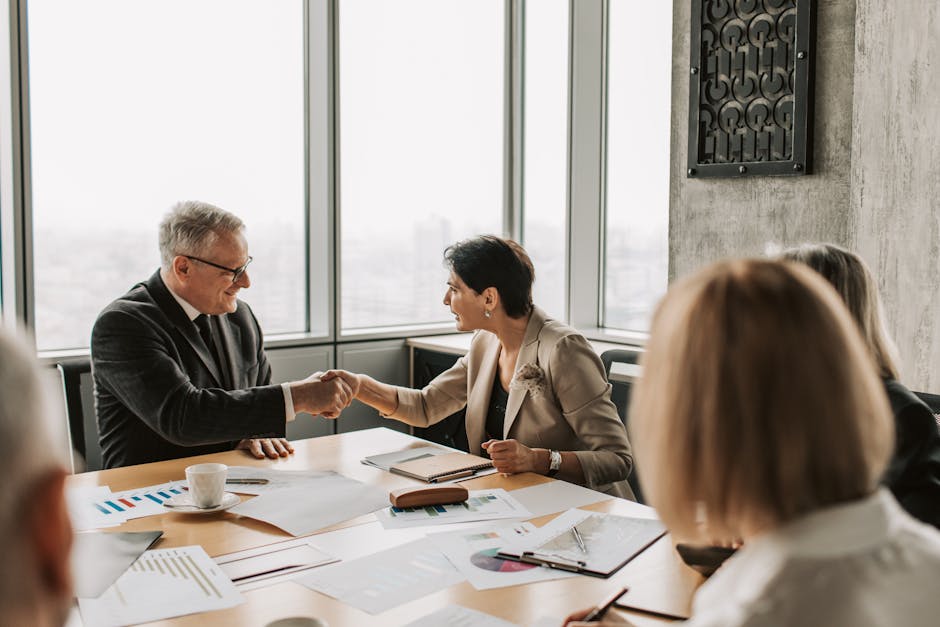 Colleagues in business attire shake hands during a formal meeting in a modern conference room.